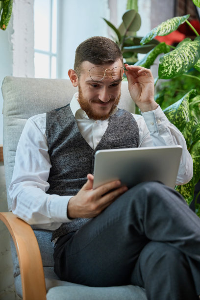 年轻放松和平的印度东部男子坐在现代客厅的沙发上，拿着平板电脑查看社交媒体，上网，订外卖，周末阅读电子书。图片下载