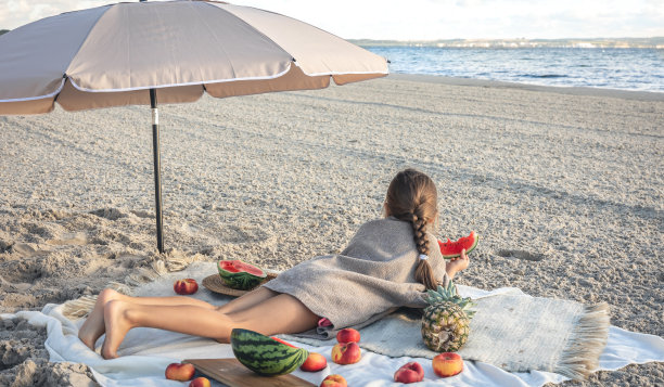 小女孩躺在沙滩上的毯子上吃水果。图片下载