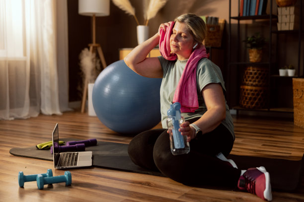 训练过程中疲惫的老妇人用毛巾擦着擦着，闭着眼睛，拿着装水的瓶子，坐在瑜伽垫上休息放松，网上瑜伽课结束后。笔记本电脑放在地板上，旁边放着哑铃图片下载