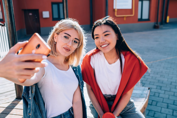 快乐的多样化朋友微笑在前面的智能手机相机，使自拍影响内容，快乐的潮女孩休息在城市设置使用蜂窝技术点击照片图片图片下载