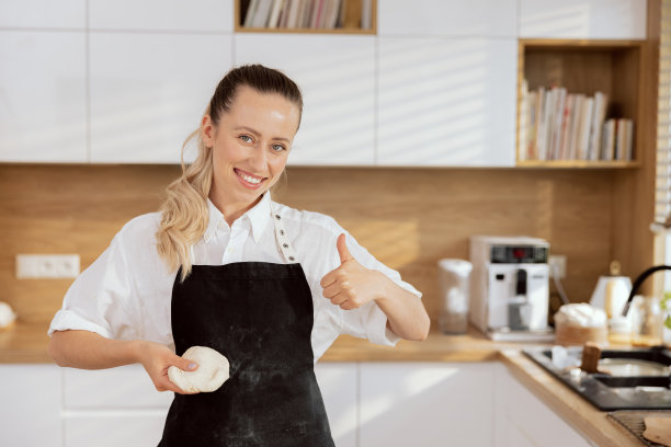 可爱的年轻女子厨师母亲，妻子穿着围裙，竖起大拇指，手捧自制面团，在现代厨房揉煮烘焙过程图片下载