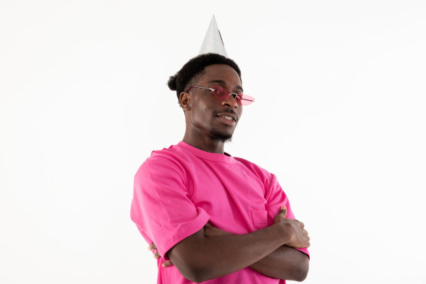 快乐快乐的非洲人看着相机严肃地戴着生日帽和粉色t恤庆祝生日摆姿势在白色背景的摄影棚里图片下载