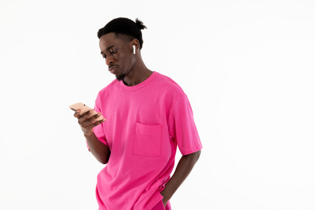 严肃的非裔美国人手里拿着手机，戴着耳机，听着音乐，严肃地看着手机屏幕，穿着粉色运动服，站在白色背景的工作室里图片下载