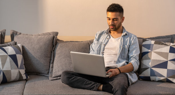 画像年轻，英俊和忙碌的男人，工作在他的笔记本电脑，坐在沙发上，穿着休闲的衣服图片下载