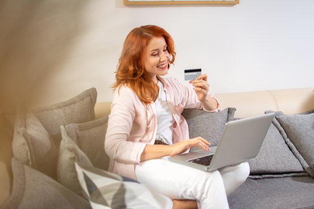 年轻快乐的姜黄色女人用笔记本电脑和手拿信用卡购物，坐在家里的沙发上。图片下载