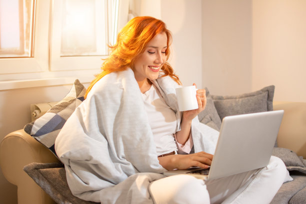 快乐的姜黄色年轻女人喝着咖啡，盖着毯子，坐在家里的沙发上用笔记本电脑工作。图片下载