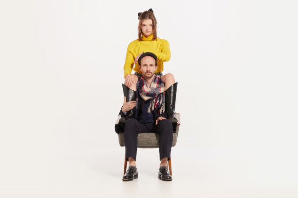 肖像野蛮的男人在秋天的外套和帽子坐在椅子上和时髦的女孩在黄色毛衣和靴子坐在男人的肩膀上图片下载