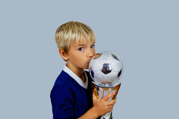在明亮的背景下，一个穿着蓝色球衣的年轻足球运动员在进球后捧着优胜者杯图片下载