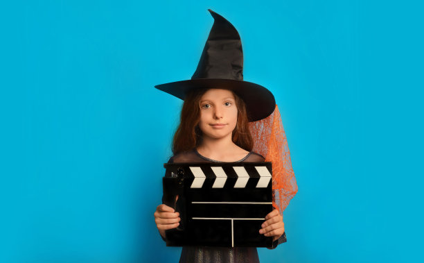 小女巫女孩戴着巫师女巫帽，手持经典的黑色电影制作隔板，看着蓝色背景上的相机。图片下载