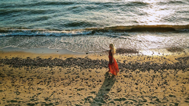 夕阳西下，身穿红色长裙的金发年轻女子走在海边图片下载
