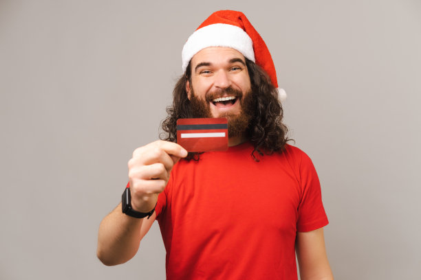 年轻惊讶的胡子男子戴着红色圣诞老人的帽子显示红色信用卡的肖像图片下载