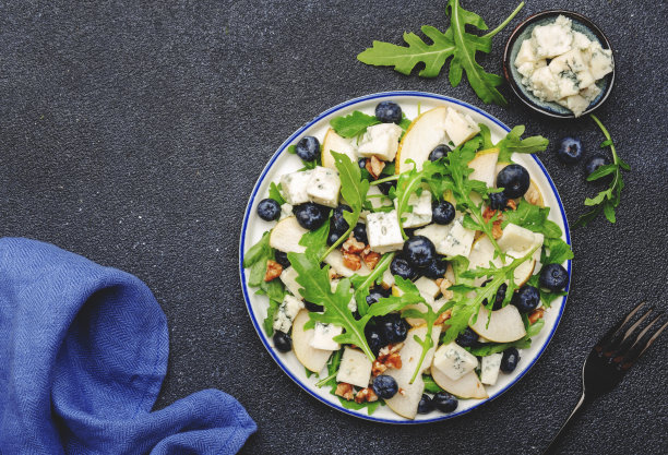 美味的芝麻菜沙拉配梨、蓝莓、羊乳干酪和核桃。黑色厨房餐桌背景，俯视图图片下载