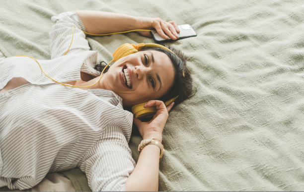 一位黑发美女戴着耳机躺在家里的床上。图片下载