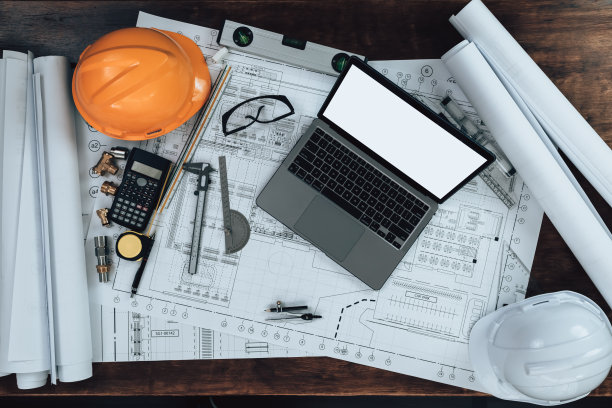 办公桌上有检查图纸的笔记本电脑，图纸上有计算器，三角尺，安全眼镜，指南针，游标卡尺。工程师，建筑师，工业和工厂概念。图片下载