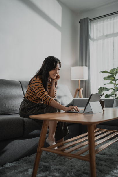 亚洲妇女开心地微笑着在平板电脑键盘上打字，在家里的客厅里工作、学习或浏览社交媒体。图片下载