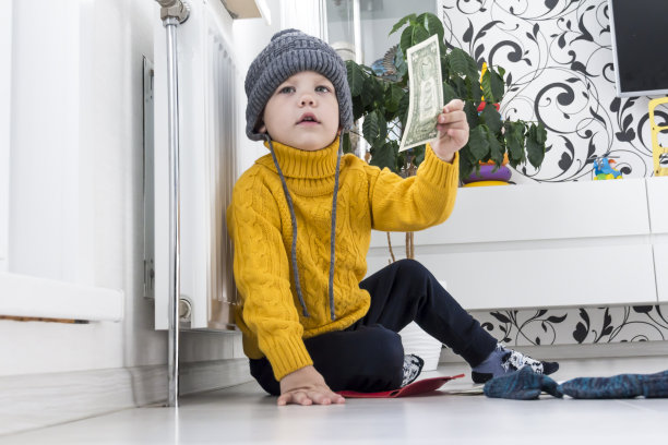 一个穿着黄色毛衣、戴着帽子的小男孩正在数钱、研究取暖费账单，旁边是一个带恒温器的加热器。图片下载