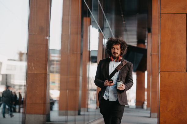时髦的商人带着皮制公文包在金融区用智能手机听着音频歌曲，戴着耳机喝着咖啡图片下载