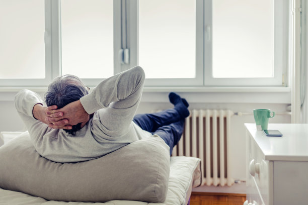 后视图一个放松的老人舒适地坐在床上，腿放在家里的暖气片上，在冬天的雨季透过大窗户。男子用加热器暖脚。图片下载