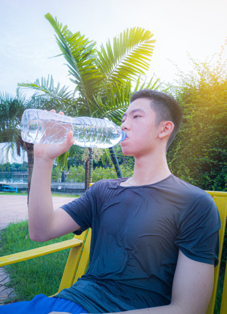 一个十几岁的年轻人坐着，放松地喝着一瓶水。早上慢跑后，汗湿了衬衫。英俊的年轻人身材匀称，肌肉发达，锻炼后能休息。图片下载