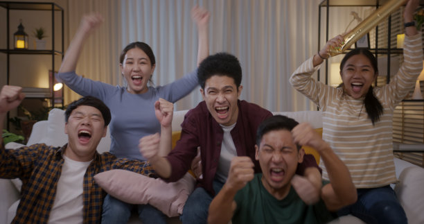 一群年轻的成年朋友男女亚洲人坐在沙发沙发欢乐的派对有趣的游戏FIFA世界杯直播电视在家里唱着歌手举着牌子跳着疯开心赢欢腾的脸。图片下载