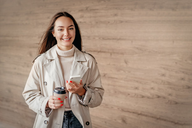 女学生面带微笑，手里拿着手机和一杯咖啡，背景是大楼的立面图片下载