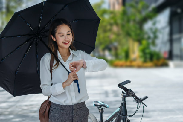 在阳光明媚的日子里，可爱的亚洲商务女性拿着黑色雨伞在户外看手表图片下载