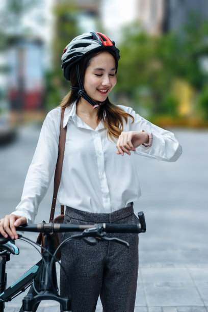 年轻的亚洲女商人戴着头盔，站在城市的街道上，骑着自行车看着手表。图片下载