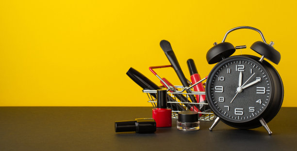 “黑色星期五”的概念。照片闹钟购物车装饰化妆品刷唇彩铅笔睫毛膏指甲油和眉毛凝胶在黑色桌面黄色的墙壁背景与copyspace图片下载