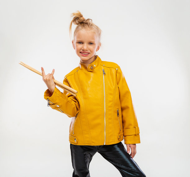 摇滚音乐概念。女音乐家，手里拿着鼓棒。穿着芥末色的皮夹克，黑色打底裤和靴子。高级时尚，与世隔绝的工作室背景图片下载