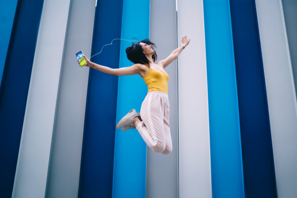 兴奋的潮女孩戴着耳机在积极的休闲时间与充满活力的音乐播客欢欣鼓舞，快乐的千禧一代女性用智能手机听有趣的媒体播放列表跳舞和跳跃图片下载