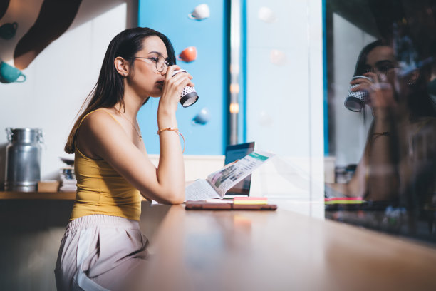 沉思的女记者戴着保护视力的光学眼镜，喝着早晨的咖啡，拿着新鲜的报纸阅读每日新闻，沉思的时髦女孩品尝咖啡因饮料图片下载