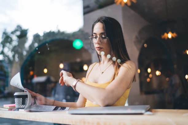 窗外的高加索女顾客坐在咖啡店等人在早上的时间，时髦的女孩戴着眼镜拿着日报，在可佩戴的手表上查看时间图片下载