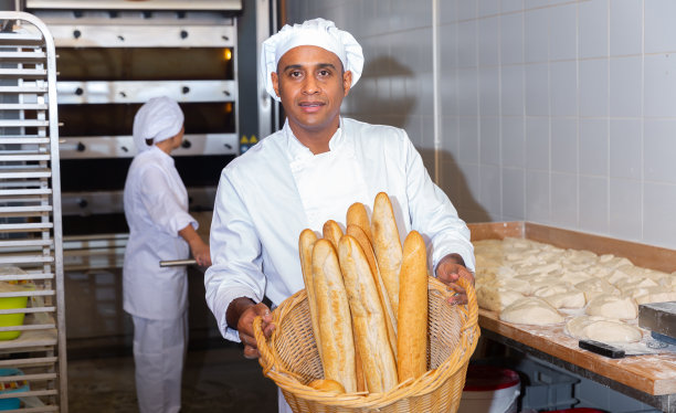 在面包店的厨房里，男面包师拿着一篮子热的法棍面包图片下载