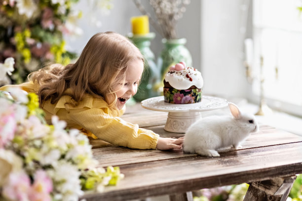 一个金发碧眼、穿着黄裙子的蹒跚学步的小女孩把手伸向桌子对面的一只活白兔。的情绪。用鲜花和复活节蛋糕装饰桌子，为复活节这个明亮的节日。中国年图片下载