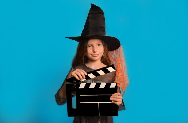 小女巫女孩在万圣节巫师服装手持经典的黑色电影制作隔板，看着相机在蓝色的背景。图片下载
