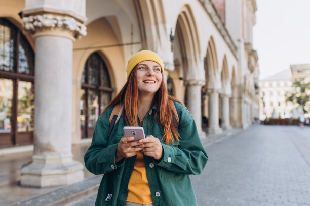 秋天的一天，美丽时髦的女人戴着帽子走在克拉科夫的市场广场上，手里拿着手机。电话沟通。城市生活方式的概念。查看社交网络，发短信。图片下载