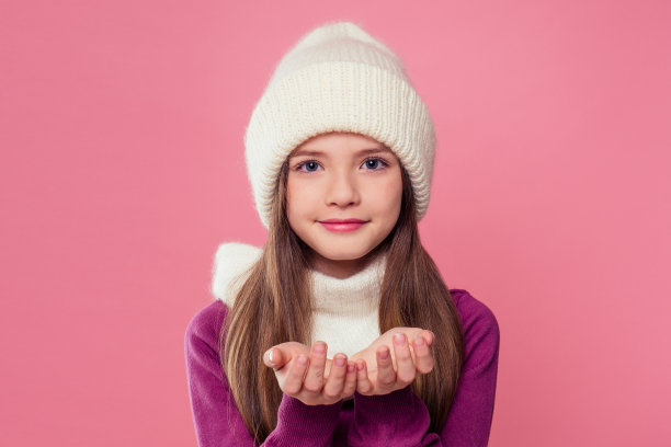 肖像可爱的小女孩在温暖的针织彩色灰色帽子，围巾和手套送吹一个吻的空气在粉红色的背景在工作室。儿童冬装秋装图片下载