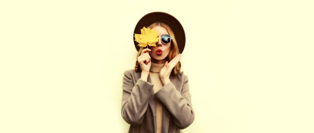 秋天的肖像，美丽的年轻女子模型吹着黄色枫叶的嘴唇与口红戴着圆帽子，白色背景的外套图片下载