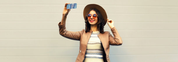 肖像快乐微笑的年轻女子用智能手机自拍戴着圆帽子，夹克在灰色背景图片下载