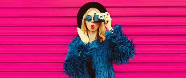 肖像时尚的金发年轻女摄影师与胶卷相机拍照和吹她的嘴唇送空吻穿着蓝色毛皮大衣，黑色圆帽在粉红色的背景图片下载