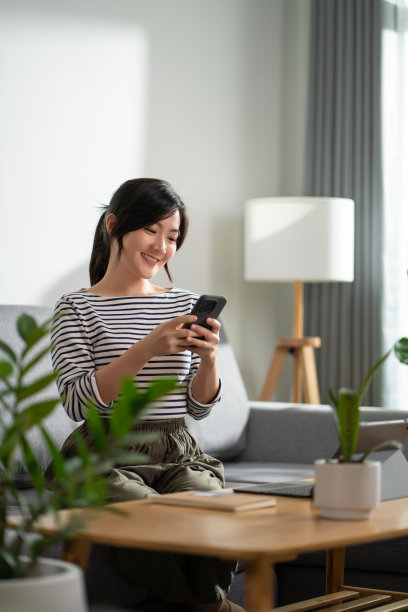 亚洲妇女快乐地微笑着坐在沙发上，在家里使用智能手机。图片下载