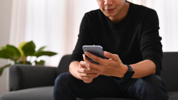 裁剪的图片中，穿着温暖的毛衣的男人在网上聊天，坐在家里的沙发上用智能手机查看社交媒体。图片下载