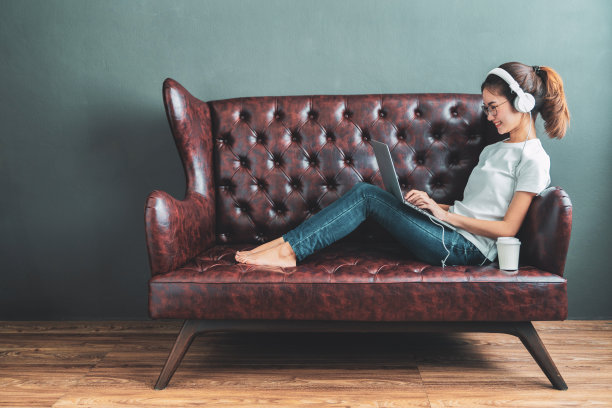 美丽年轻的亚洲女人戴着耳机在沙发上放松。她正在用笔记本电脑或平板电脑听音乐。放松和休闲的概念图片下载