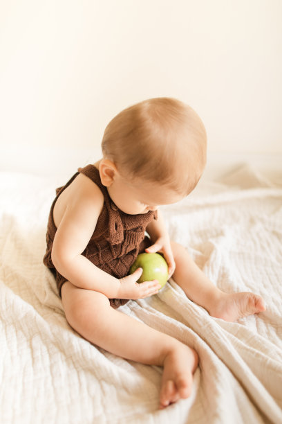 2022年秋天，一个9个月大的大流行男婴穿着可持续制作的棕色针织紧身衣，在一个新鲜的绿色史密斯奶奶苹果上玩耍和长牙，坐在家里地板上的奶油色薄纱毯子上图片下载