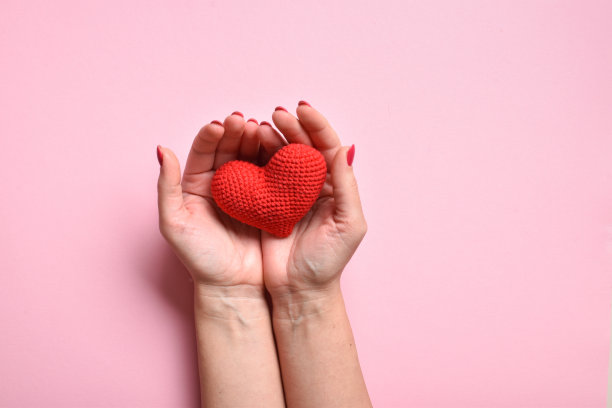 粉红色背景上，一个有着漂亮指甲的女孩手里拿着一颗红色编织的心。爱。器官捐赠。怀孕。情人节。副本的空间。平铺，俯视图图片下载
