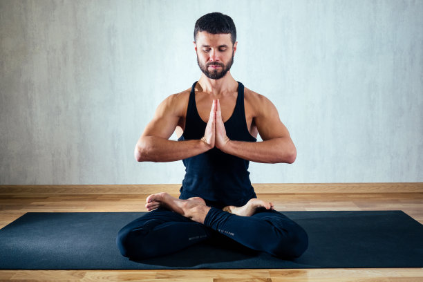 灰色背景下，一名身穿深色运动服的男子正以莲花姿练习瑜伽。在瑜伽垫上做瑜伽体式。身体的集中和占有的概念图片下载
