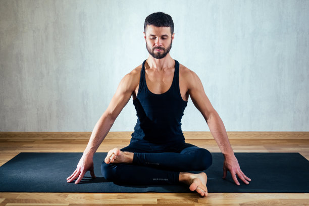 灰色背景下，一名身穿深色运动服的男子正以莲花姿练习瑜伽。在瑜伽垫上做瑜伽体式。身体的集中和占有的概念图片下载