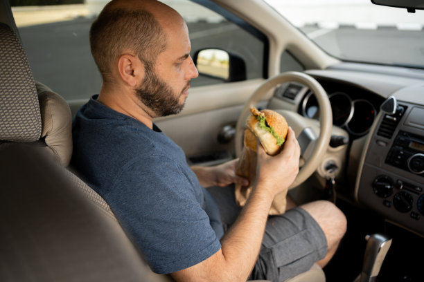 男人一边开车一边吃汉堡包。饥饿的司机一边开车一边在车流中吃东西图片下载