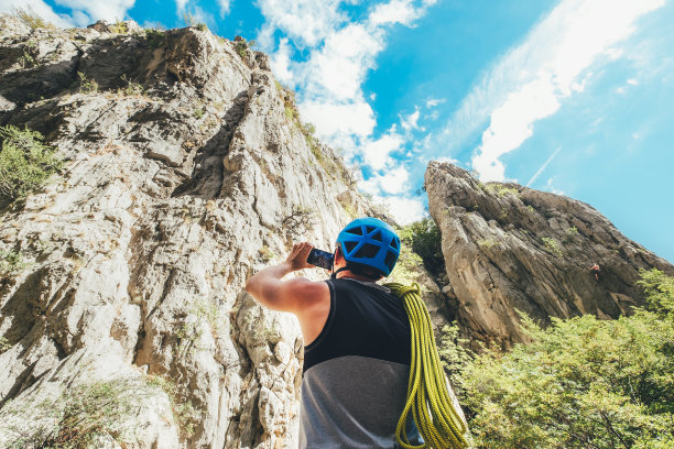 在Paklenica的岩壁之间，戴着防护头盔、肩上有攀岩绳的登山者正在用现代智能手机拍摄悬崖照片。积极的极限运动时间消费理念图片下载
