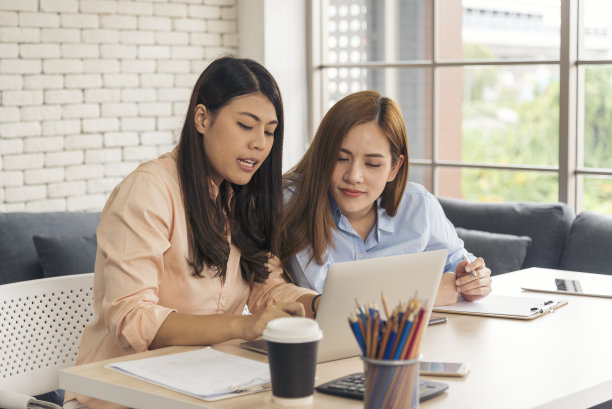 两个女商人在公司办公桌旁用笔记本电脑开会。两个年轻的女性自由职业阅读金融图表策划分析市场数据。亚洲人团队工作办公室公司。图片下载